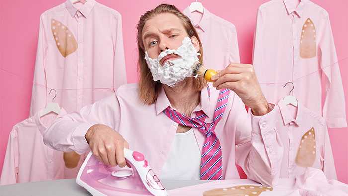 Guy simultaneously shaving and ironing surrounded by burned shirts