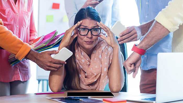 Overwhelmed job candidate sits at a desk surrounded by co-workers asking questions, passing off phone calls, and pointing out the time