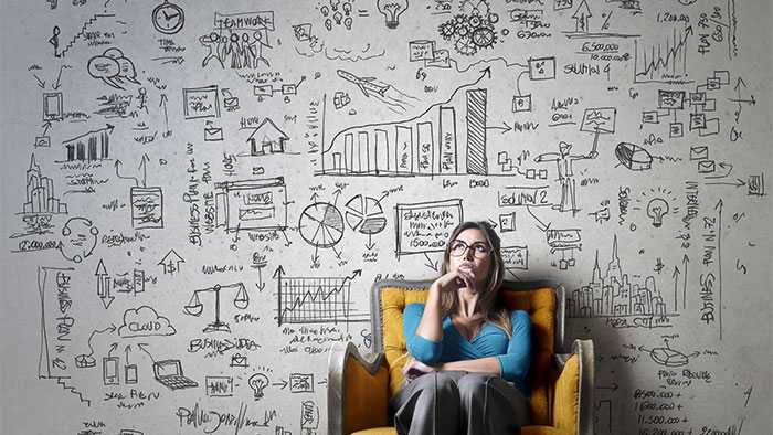 A woman sits in a chair looking up in thought in front of a wall covered with hundreds of charts and concepts that she's managing